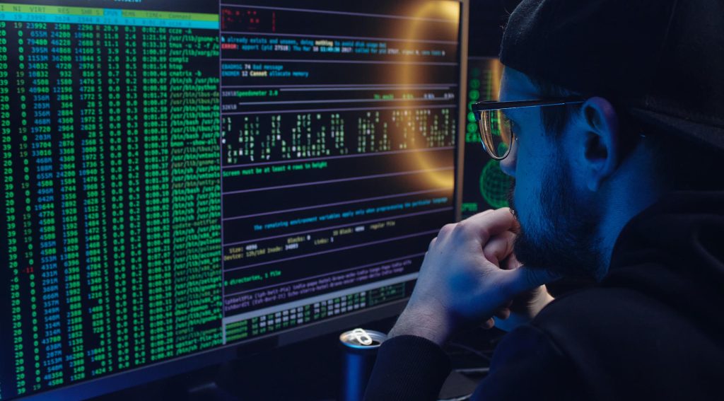 Cybercriminal looking at lines of code on two computer screens in a darkened room